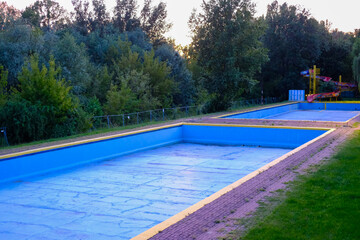 Empty outdoor swimming pool with bright blue walls and yellow edges, set in a green park environment at dusk.