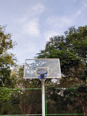 An outdoor basketball hoop with a transparent backboard, surrounded by lush green trees under a bright afternoon sky