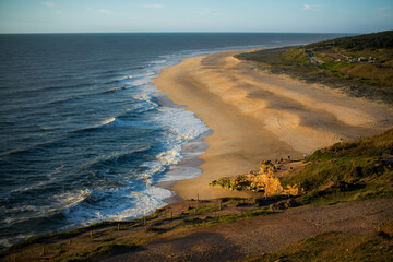 The ocean beach in Nazaré rests under calm weather, with gentle waves lapping the shore and soft light reflecting on the water. The scene feels peaceful, open, and quietly vast.