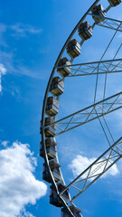Partial view of a Ferris wheel against a bright blue sky with scattered clouds. The modern cabins are visible in detail.