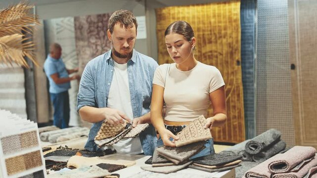  Couple of young woman and man buyers choosing soft carpet samples in building store