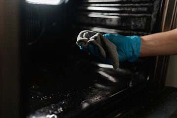 Professional cleaner worker wearing blue rubber gloves wiping inside of oven with gray rag,...