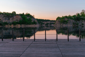 Wide wooden platform with metal railing overlooking a calm lake at sunset. Forested limestone...