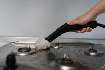 Male hand using steam cleaner with cloth attachment sanitizing grimy gas stove surface,...