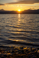 Beautiful sunset over the mountains above the lake. Reflection of sun rays in the water in a sea bay with trees