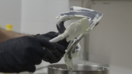 Professional pastry chef wearing gloves carefully scraping sticky dough from mixer paddle using...