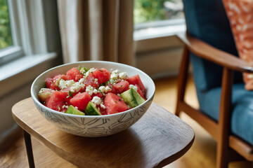 Fresh watermelon salad served in a decorative bowl on a wooden table next to a cozy chair by the window