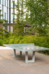 A sleek metallic table tennis setup on a paved surface, surrounded by plants and trees. A modern...