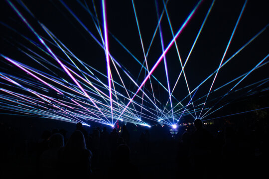 Intersecting blue, pink, and white laser beams light up the dark night as silhouettes of people watch, suggesting a public event or performance.