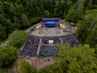 An aerial view of an outdoor amphitheater surrounded by tall trees, featuring a stage with blue...