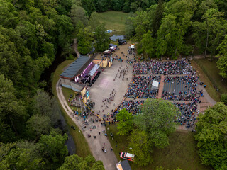 An outdoor amphitheater hosts a performance with a seated audience, surrounded by trees, pathways,...