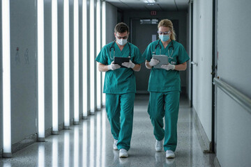 Young adult Caucasian man and young adult Caucasian woman doctors walking down hospital corridor holding digital tablets and wearing medical masks and gloves, discussing patient information