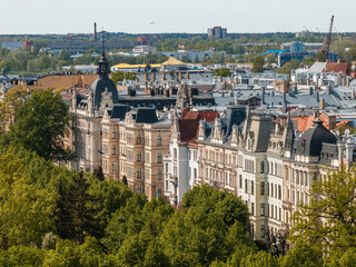 Row of ornate historic buildings with steep roofs, trees in the foreground, and modern buildings in...