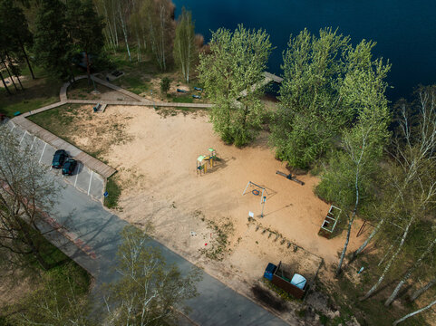 A sandy area near water with a playground, wooden dock, trees, pathways, benches, a paved parking lot, and a mix of open and wooded spaces. - Powered by Adobe