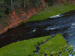 A person in a blue jacket kayaks on a dark river bordered by green grass and a red sandstone cliff...