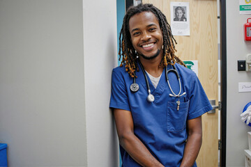 Portrait of young adult Black man doctor smiling at camera, standing in medical facility with stethoscope around neck, displaying friendly and approachable demeanor