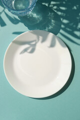 Empty white plate with glass of water on the blue background with palm leaves shadows. Summer table...