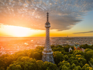 At sunrise, the Petrin Lookout Tower in Prague rises above a lush park, bathed in golden light,...
