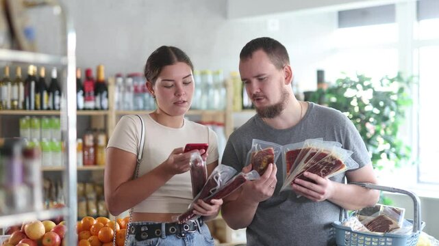 In minimarket, couple buyers inspects hamon and dried sausage, photographs scan QR code on product label. Visitor places discount on purchase in stores app, checks product reviews in mobile app