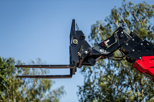 industrial forklift attachment on hydraulic crane arm with metal prongs lifting machinery in construction site against blue sky and green trees heavy equipment closeup for cargo transport and logistic