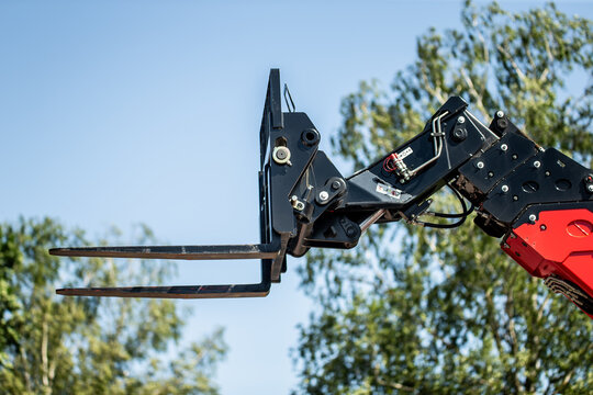 close up of hydraulic forklift attachment on red construction vehicle with lifting forks extended against clear blue sky and green trees, industrial machinery for loading transport and heavy lifting t