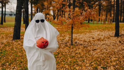 Woman in white sheet holding jack o'lantern against autumn forest background. Halloween costume. 