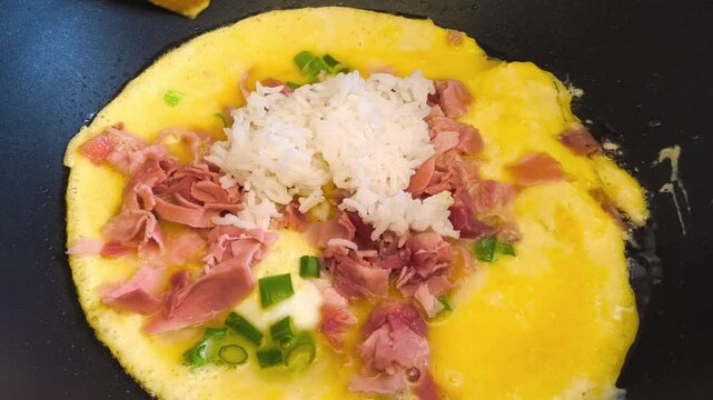 Close-up view of egg mixture sizzling in a hot frying pan on a stove with high heat, preparing a soft Asian Japanese omelette with rice, ham, and sliced green onions, for brunch, breakfast, lunch meal