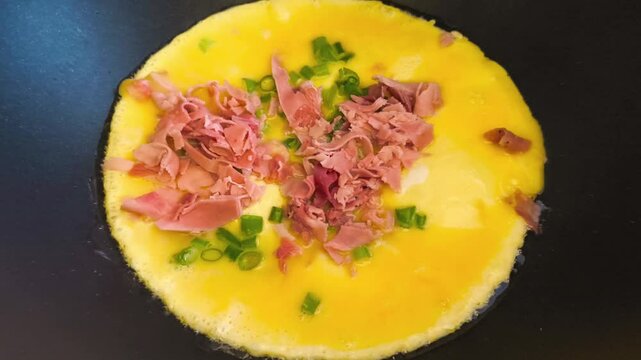 Close-up view of cooking an omelette as ham and sliced green onion egg mixture is poured into a hot oiled non-stick pan on high heat, making a soft Asian Japanese omelette in a kitchen cooking process