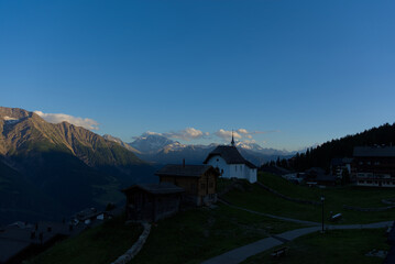 A serene church with a cross-topped steeple stands silhouetted against snow-capped mountains and a...