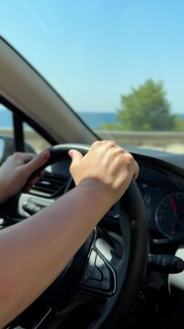 Close-up of hands on a steering wheel while driving along a sunny coastal road. The sea and trees blur in the background, evoking a peaceful summer vibe and freedom of travel