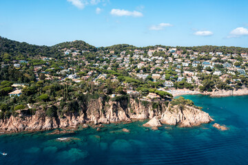 An aerial view of Blanes showcasing a beautiful bay with clear turquoise water, rocky cliffs, and numerous white villas nestled among green trees on the hillsides