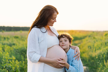 Obraz premium A pregnant woman stands in a lush green field, gently holding her baby bump. Her son leans against her, both sharing a tender moment amidst tall grass and a warm sunset.
