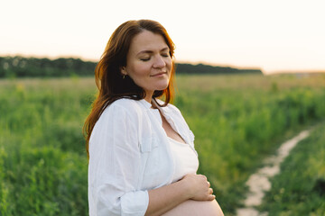 A pregnant woman stands in a green field. She gazes lovingly at her baby bump as the sun sets behind her, creating a serene atmosphere.
