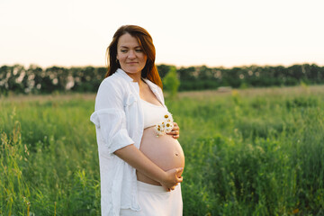 A pregnant woman stands in a green field. She gazes lovingly at her baby bump as the sun sets behind her, creating a serene atmosphere.