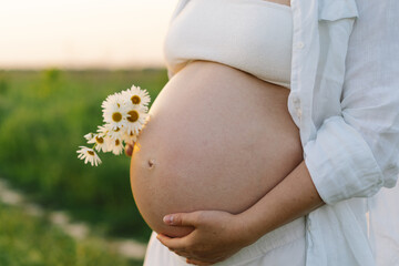 A pregnant woman stands in a green field. She gazes lovingly at her baby bump as the sun sets behind her, creating a serene atmosphere.