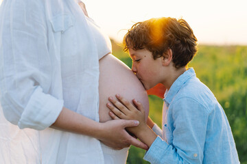 A pregnant woman stands in a lush green field, gently holding her baby bump. Her son leans against her, both sharing a tender moment amidst tall grass and a warm sunset.