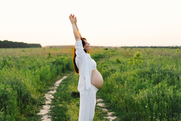 A pregnant woman stands in a green field. She gazes lovingly at her baby bump as the sun sets behind her, creating a serene atmosphere.