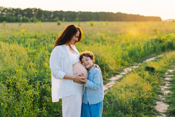 A pregnant woman stands in a lush green field, gently holding her baby bump. Her son leans against her, both sharing a tender moment amidst tall grass and a warm sunset.