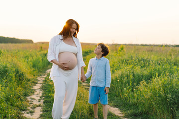 A pregnant woman stands in a lush green field, gently holding her baby bump. Her son leans against her, both sharing a tender moment amidst tall grass and a warm sunset.
