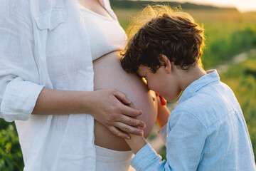 A pregnant woman stands in a lush green field, gently holding her baby bump. Her son leans against her, both sharing a tender moment amidst tall grass and a warm sunset.