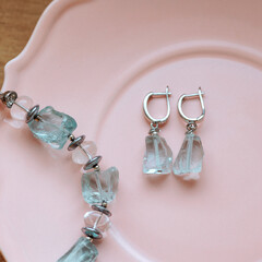 Chunky silver earrings with raw crystals and matching bracelet with clear and aqua stones on pink ceramic plate, wooden background. Concepts: handmade jewelry, accessories, elegance, gift idea.