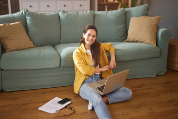 A young woman enjoys a relaxed atmosphere in her home office as she works on her laptop, seated...