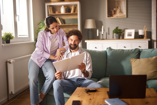 A couple sits on a couch in their living room, engaged in a discussion about life insurance while reviewing documents. Natural light fills the room, creating a warm atmosphere.