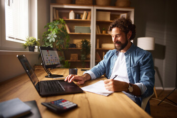 A man focuses on his tasks at a stylish home office, writing notes while using a laptop and surrounded by greenery and wooden shelves filled with decorative items.
