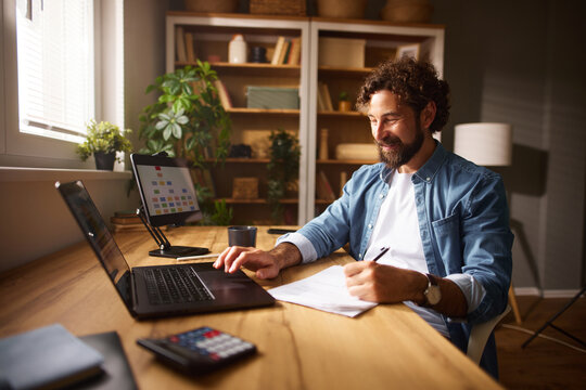 A man focuses on his tasks at a stylish home office, writing notes while using a laptop and surrounded by greenery and wooden shelves filled with decorative items.