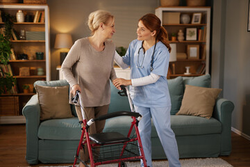 A home health aide assists an elderly woman with her walker in her living room. The healthcare worker, wearing scrubs and a stethoscope, helps the senior citizen with mobility.