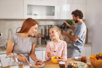 A mother smiles at her daughter while spreading jam on toast, and the girl enjoys a bowl of fruit....