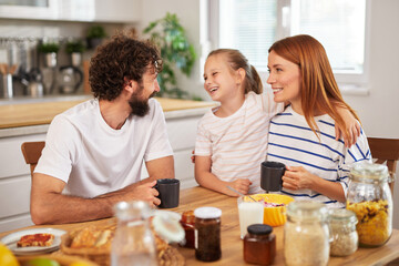 A family engages in cheerful conversation during breakfast in their kitchen. The daughter shares a playful moment with her parents while enjoying coffee and homemade food.