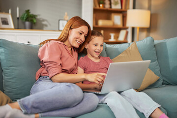 A smiling mother and her daughter sit together on a comfortable couch, engaging with a laptop. They share a joyful moment as they explore online content in their inviting living room.