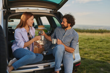 Two friends are seated at the back of their car enjoying a cozy picnic in a beautiful outdoor location. They are sharing warm drinks while chatting happily in the golden light.